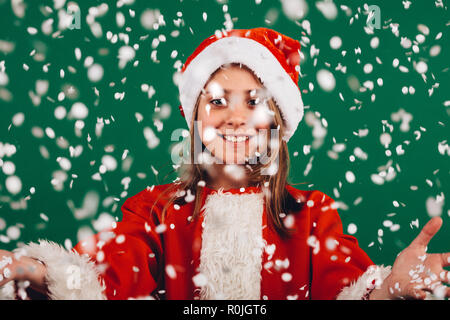 Lächelnde Mädchen in santa claus Kleid vor grünem Hintergrund mit künstlichem Schnee Flocken fallen alle um. Porträt eines lächelnden Mädchen mit Op Stockfoto