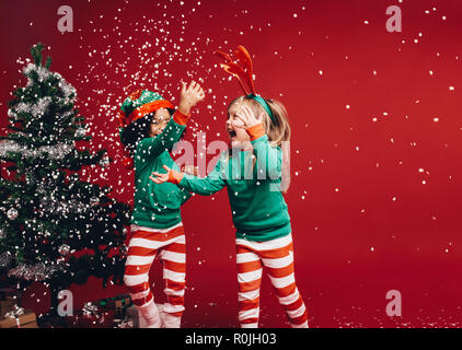 Kinder spielen mit künstlichem Schnee Flocken fallen in der Nähe einen Weihnachtsbaum. Kleine Mädchen in Weihnachten Kostüme Spaß spielen neben einer dekorierten Chris Stockfoto