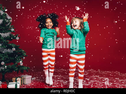 Kleine Mädchen in Weihnachten kostüm Spaß und Springen neben einem geschmückten Weihnachtsbaum. Zwei Kinder spielen mit fallender künstliche Schneeflocken in der Nähe von Stockfoto