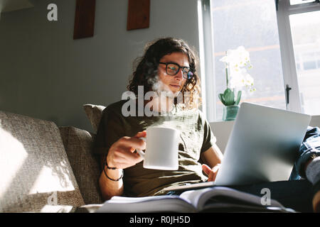 Student Studium zu Hause sitzen auf der Couch neben einem Fenster. Junge Mann hält eine Tasse Kaffee studieren auf dem Laptop. Stockfoto