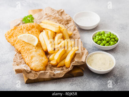 Traditionelle britische Fisch und Chips mit Sauce Tartar auf Schneidebrett und grünen Erbsen auf weißen Stein Hintergrund. Zitronenscheibe und Salz Stockfoto