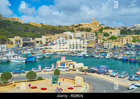 GHAJNSIELEM, MALTA - 15. JUNI 2018: Hafen von Mgarr mit alten Fischerbooten ist von hohen Hügeln mit privaten Cottages und Lourdes Kapelle auf t umgeben Stockfoto