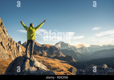 Sportliche Mann stand auf dem Stein mit erhobenen Armen gegen die Berge bei Sonnenuntergang. Glücklich, junger Mann, Felsen, Wald und blauem Himmel in Dolomit Stockfoto