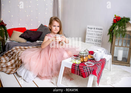 Hübsches kleines Mädchen im Alter von 4 Jahren in ein rosa Kleid. Kind in der Weihnachtszeit Zimmer mit einem Bett, Essen, Süßigkeiten, Schokolade, Kekse und Tee trinken, Fütterung ted Stockfoto