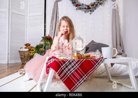 Hübsches kleines Mädchen im Alter von 4 Jahren in ein rosa Kleid. Kind in der Weihnachtszeit Zimmer mit einem Bett, Essen, Süßigkeiten, Schokolade, Kekse und Tee trinken, Fütterung ted Stockfoto