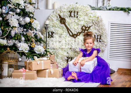 Hübsches kleines Mädchen 4 Jahre alt in einem blauen Kleid. Baby in Weihnachten Zimmer mit Teddybär, big Clock, Weihnachtsbaum, braune Sessel, Kerzen und Geschenke. Stockfoto