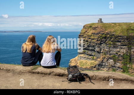 Zwei junge weibliche Touristen sitzen in Richtung O'Brien's Tower und die Aran Inseln auf den Klippen von Moher an der Atlantischen Westküste Irlands suchen Stockfoto