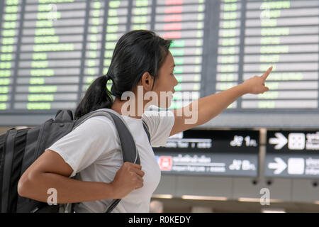 Reisen Frau mit einem Rucksack auf dem Rücken, die ein Schild mit den Flugverbindungen. Die asiatische Mädchen mit dem Gepäck ist unter Vorstand am Flughafen. Stockfoto