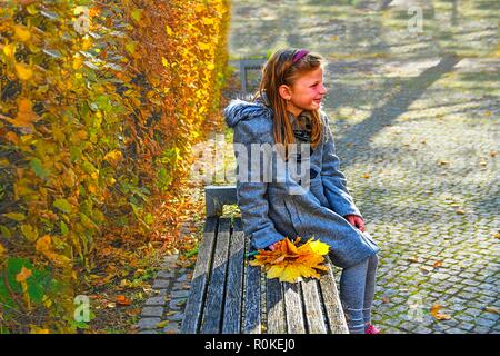 Kleines Mädchen tragen Retro Rock und sitzt auf der Bank im Park im Herbst. Kleines Mädchen hält bunte Blätter im Herbst. Herbst Konzept Stockfoto