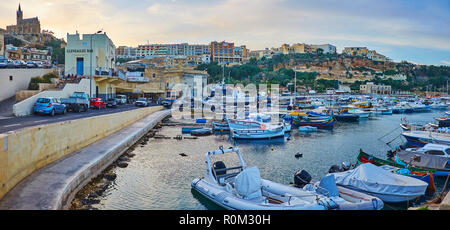 GHAJNSIELEM, MALTA - 15. Juni 2018: Das stadtbild von der Hafen von Mgarr mit Fischerbooten, steile Hügel, Lourdes Kapelle und Wohngebiete Stockfoto