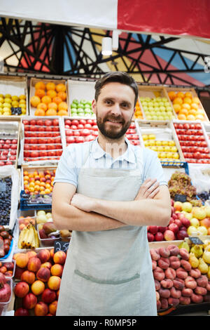 Inhalt hübscher junger grocery Retailer mit Bart tragen Schürze stehend gegen frische Lebensmittel Regale und lächelnd an der Kamera während der Überfahrt Arme auf ches Stockfoto