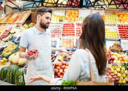 Verwirrt Missverständnis junge männliche Lebensmittelhändler in Schürze halten Behälter mit Erdbeeren zuckte mit den Schultern, während im Gespräch mit Kunden in Farmers Market Stockfoto