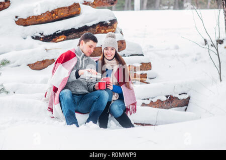 Junge liebende Paar in blauen Pullover sitzen mit roten Kaffeetassen auf dem Brennholz im Winter Wald angezogen Stockfoto