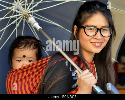 Jungen nordöstlichen indischen Monpa Frau mit modernen Brillen trägt ihr baby boy in einem traditionellen Babytragetuch unter einen Schirm auf dem Rücken. Stockfoto