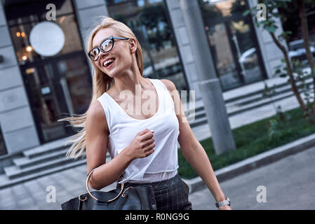 Junge hübsche Geschäftsfrau in Sonnenbrille lächelnd und Wandern in der Nähe von Business Gebäude. Sie ist ist ein eilig zu der Sitzung mit ihren Partnern. Stockfoto