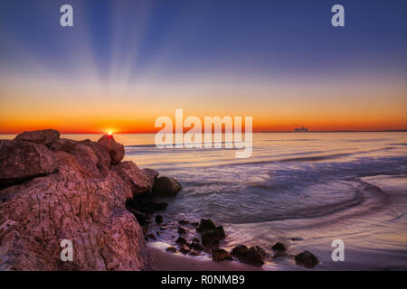 Eine tiefe und farbenfrohen Sonnenaufgang über Felsen und Sandstrand Meer mit Blau, Rot, Orange und Gelb, und ein Schiff in die Ferne angedockt an einem Stockfoto
