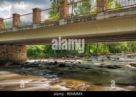 Weich fließende Fluss gehen unter einen Stein und Stahl Brücke mit der Sonne reflektiert das Wasser und Felsen und Moody Wolken im Himmel Stockfoto