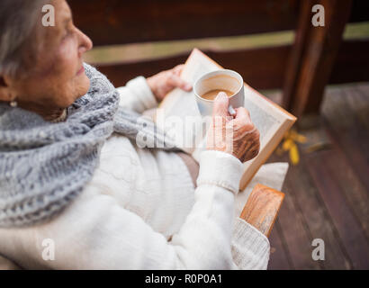 Eine ältere Frau liest Buch draußen auf einer Terrasse an einem sonnigen Tag im Herbst. Stockfoto