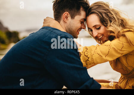 Seitenansicht eines jungen Paares zusammen sitzen, ihre Köpfe zu berühren. Romantisches Paar auf ein Datum in der Nähe von einem See jedes andere Holding sitzen. Stockfoto