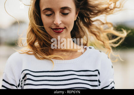 Nahaufnahme von einer jungen Frau, die draußen mit ihrem Haar fliegen. Porträt einer Frau mit einem angenehmen Lächeln stand im Freien. Stockfoto