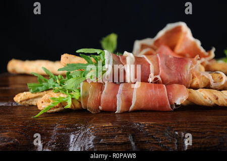 Grissini - Brot Sticks mit Parmesan, umwickelt mit einem Stück Schinken und Rucola. Italienische Gericht mit Antipasti auf einem Holztisch. Stockfoto