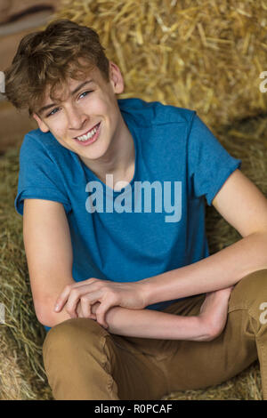 Junger Mann glücklich lächelnde Lachende männliche junge Teenager sitzen auf Strohballen in einer Scheune Stockfoto