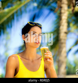 Junge Frau mit gelben Badeanzug mit einem Glas Bier, Palmen Hintergrund, Guadeloupe, Französisch Westindien, Stockfoto