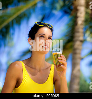 Junge Frau mit gelben Badeanzug mit einem Glas Bier, Palmen Hintergrund, Guadeloupe, Französisch Westindien, Stockfoto