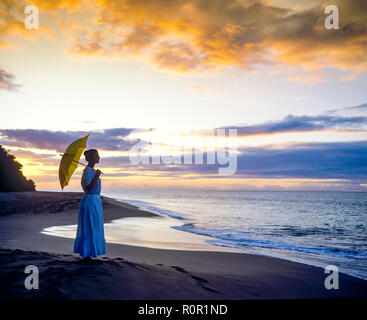 Junge Frau Silhouette mit Kleid und Sonnenschirm mit Blick auf das Karibische Meer bei Sonnenuntergang, tropischen Strand, Guadeloupe, Französisch Westindien, Stockfoto