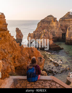 Weibliche Wanderer sitzt auf Mauer aus Stein und blickt über Felsen im Meer, Algarve, felsige Küste, Ponta da Piedade, Lagos, Portugal Stockfoto