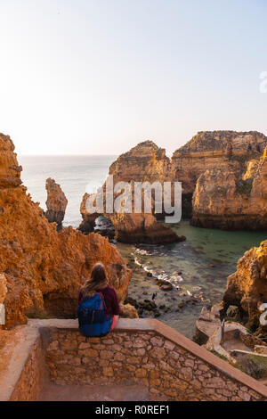 Weibliche Wanderer sitzt auf Mauer aus Stein und blickt über Felsen im Meer, Algarve, felsige Küste, Ponta da Piedade, Lagos, Portugal Stockfoto