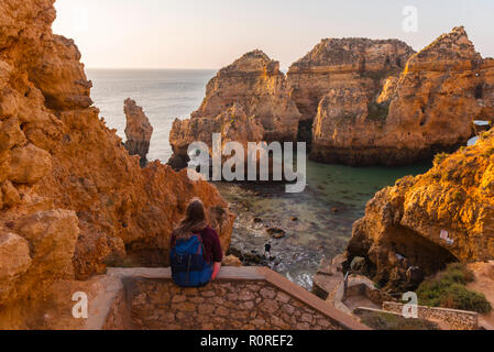 Weibliche Wanderer sitzt auf Mauer aus Stein und blickt über Felsen im Meer, Algarve, felsige Küste, Ponta da Piedade, Lagos, Portugal Stockfoto