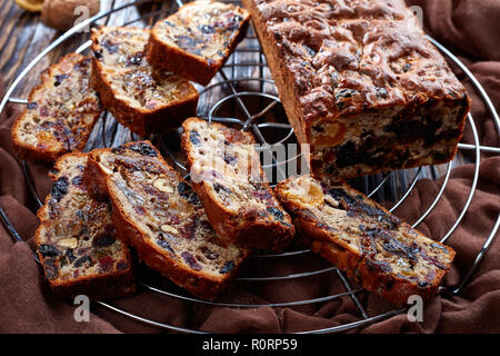 Frisch gebackene getrockneten Früchten reichen Kuchen auf einem Draht Kuchen stand mit braunem Stoff, Zimtstangen, getrocknete Aprikosen und Datum Früchte auf einem rustikalen Holztisch, Stockfoto