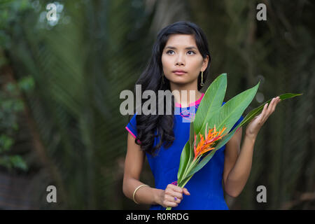 Die Hälfte Körper geschossen von einem jungen Hübschen vietnamesische Frau in Blau Ao Dai Kleid Holding orange Blüten mit grossen Blättern, Nachschlagen. Stockfoto