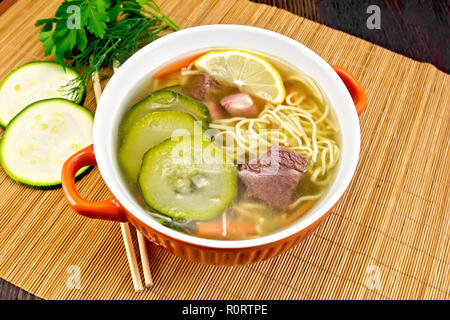 Suppe mit Zucchini, Rindfleisch, Schinken, Zitrone und Nudeln in eine Schüssel geben, Petersilie und Dill auf einem Bambus Serviette gegen einen dunklen Holz- Board Stockfoto
