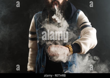 Junger Mann mit Bart werfen eine Wolke aus Dampf. Schwarzen Hintergrund. Vaping eine elektronische Zigarette mit viel Rauch Stockfoto