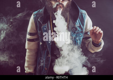 Junger Mann mit Bart werfen eine Wolke aus Dampf. Schwarzen Hintergrund. Vaping eine elektronische Zigarette mit viel Rauch Stockfoto