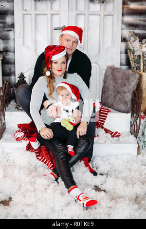 Portrait von Happy Family im roten Nikolausmützen auf Treppen mit Schnee in Füßen umarmten sitzen. Weihnachten und Familie Konzept. Stockfoto