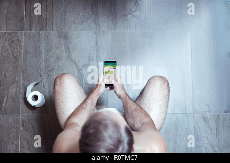 Blonde Mann mit seinem Smartphone während auf der Toilette sitzen Stockfoto
