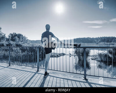 Mann stretching Beine, bevor sie nach draußen gehen in sonniger Morgen laufen. Aufwärmen der Muskeln. Im freien Training auf See brücke. Stockfoto