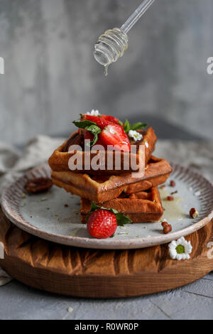 Belgische Waffeln mit Erdbeeren und Honig am grauen Platte auf Holz Brett bei Bray Hintergrund, Kamille in der Nähe von Stockfoto