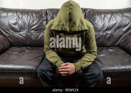 Mann im dunkel Grün hoodie mit unkenntlich Gesicht sitzen auf broun Ledersofa Stockfoto