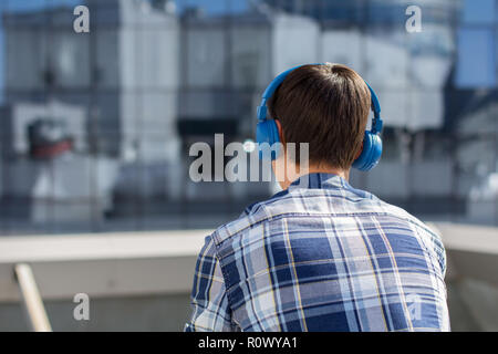 Rückansicht des jungen Mannes in kariertem Hemd mit drahtlosen Kopfhörern im Stadtbild. Kerl Hören von Musik über Kopfhörer vor moderne Build Stockfoto