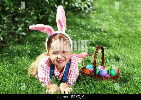 Ein fröhliches kleines Mädchen Vorschüler im bunny Ohren gekleidet liegt auf dem Rasen mit einem Korb von bemalten Ostereier. Frohe Ostern Stockfoto