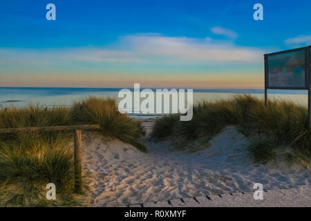 Ein Strand Eingang durch die Dünen, Gräser und feinen Sand von Fehmarn in Deutschland in der Abenddämmerung. Tolle Aussicht auf die Ostsee und die bunten ... Stockfoto