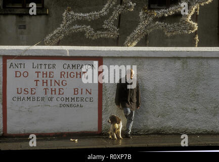 Ein Mann steht untätig und lehnt sich an eine Wand neben einem Anti-Wurf-Kampagnenschild. Sein Spaniel-Hund sitzt an seiner Seite. Über ihnen sind Weihnachtsdekorationen. Donegal, Irland Stockfoto