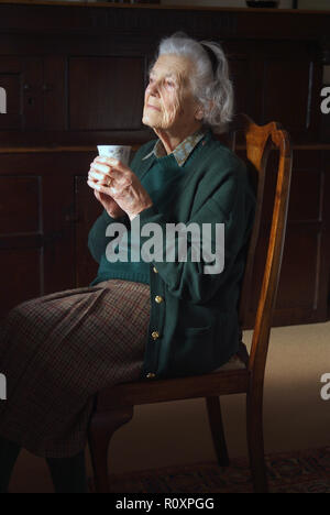 Porträt der alten Dame sitzen auf hohen Mahagoni Stuhl. Sie hält eine Tasse Kaffee und blickt zufrieden auf das Licht eines Fensters. Sie trägt ein Grün Strickjacke und braunem Tweed Rock. Ihr Haar ist weiß und ist mit einem schwarzen Band gebunden Stockfoto