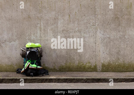 Wanderrucksack mit grünem Tuch, Bratpfanne, Schlafsack und grünen Laufschuhe stehen auf einer Straße vor einer grauen Wand Stockfoto
