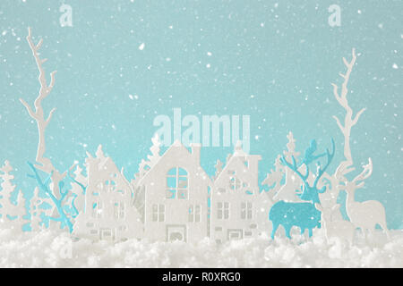 Zauberhafte Weihnachten Papier schneiden Winter Hintergrund Landschaft mit Häusern, Bäumen, Rotwild und Schnee vor Pastell-blaue Hintergrund Stockfoto