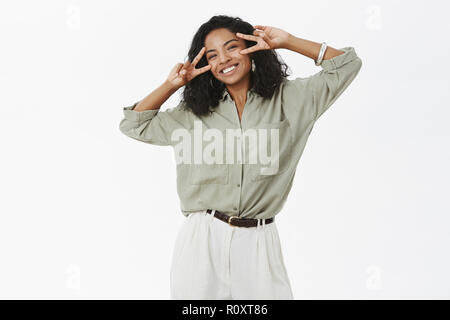 Glück ist in der Nähe. Portrait von sorglosen charmante und optimistische junge afrikanische amerikanische erfolgreiche Frauen in Bluse und Hose mit Tattoo, Frieden Zeichen über Augen und freudig lächelnd Stockfoto